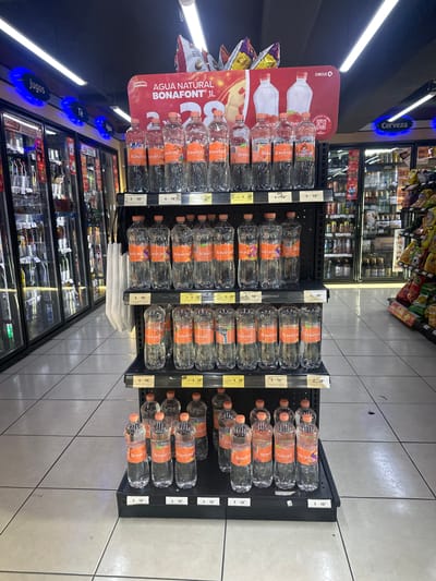 A medium-shot, high-angle photograph shows the interior of a Circle K store, likely in Jesús del Monte, Mexico, showcasing a display of "Bonafont" bottled water. Three black shelving units stand in a row, each filled with multiple bottles of the water. The bottles have orange labels. Price tags are visible next to each shelf level, with some marked at "$18" and others at "2x $28".
A red promotional sign for "Bonafont" is attached to the leftmost shelving unit. The sign features images of the water bottles and text in Spanish, including "AGUA NATURAL BONAFONT® 1L." Beneath the water display, cooler units filled with various beverages can be seen. The blue "Jugos," "Té" and "Cerveza" signage is above the display.
The store's tiled floor is visible, providing a clear, organized background. In the distance, other shelves filled with food and beverage products are present.
