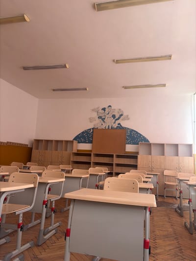 The image depicts an empty classroom, likely in a school in Vratsa, Bulgaria. The room features rows of light-colored desks with grey metal frames and red accents, arranged facing a whiteboard or projection screen that is not fully visible. The floor is made of polished wooden parquet. Along the back wall, there are multiple wooden storage units with open shelves and closed cabinets. Above the storage units, a mural or painting adorns the wall, featuring stylized figures and birds. Fluorescent light fixtures are mounted on the ceiling. The overall impression is of a clean and orderly, yet unoccupied, educational space.