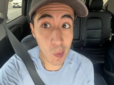 A medium-shot, front-facing selfie taken from inside a vehicle shows a person wearing a grey baseball cap and a light blue, textured t-shirt. The person is looking upwards and to their left, with their lips pursed. The background of the image reveals the interior of the car, including black leather seats and a seatbelt. Outside the vehicle, through the window, another white car is visible on a paved road. The person's face is the primary focus, with prominent eyebrows and freckles visible. A silver chain necklace is also apparent. The scene appears to be set in a city, with the presence of vehicles and infrastructure suggesting an urban environment. It is not possible to determine the specific location within Monterrey, Mexico based on the provided image.