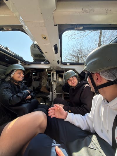 The image depicts three individuals seated within the interior compartment of a military armored vehicle in Vila Real, Portugal, engaged in an interaction. A fourth individual is partially visible further inside the vehicle.
On the left, a female with dark hair wears a black leather jacket and a dark green military helmet over a white hairnet. She is smiling and looking towards the right, with her arms crossed. A black handbag is positioned next to her. In the center, a male with dark hair wears a dark brown hoodie and a dark green military helmet. He is also smiling and looking towards the right. Partially visible in the immediate foreground on the right, a male wears a white hoodie and a dark green military helmet over a white hairnet. His right hand is open, appearing to gesture during a conversation. A black bag with a "Guess" logo is visible below his arm.
In the background, the lower torso and legs of a fourth person are visible, wearing camouflage military trousers and light brown boots. The vehicle's interior features light-colored metal panels, structural supports, and ventilation openings. Through the open windows or hatches, a clear blue sky and bare tree branches are visible, indicating an outdoor, daytime setting. Natural light illuminates the scene from above and the sides.