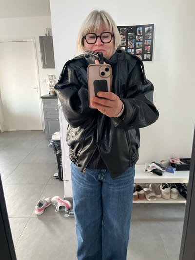 A woman with short blonde hair and glasses is taking a selfie in a mirror. She is wearing a black leather jacket and blue jeans. The background includes a white wall with a framed collage, a shoe rack filled with shoes, a kitchen area with gray cabinets and a white door, and gray tile flooring. Two pairs of shoes are on the floor. The location is likely inside a residence in Chambœuf, France, given the provided information.