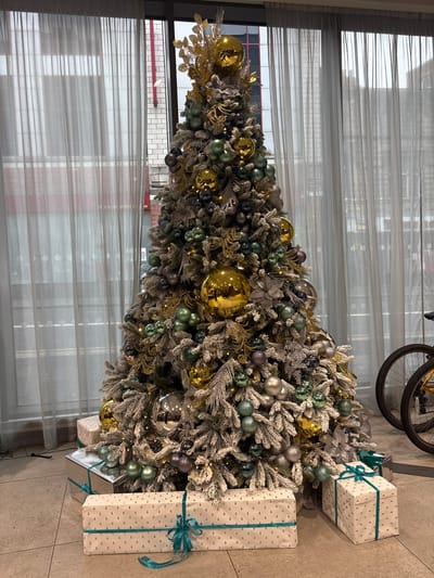 An indoor vertical shot depicts a large, frosted artificial Christmas tree centrally placed on a tiled floor. The tree is heavily decorated with an assortment of spherical baubles in metallic gold, various shades of green, silver, and muted purple, interspersed with gold glittered foliage, silver poinsettia-like flowers, and warm white string lights. At the base of the tree are several wrapped gifts: a prominent long rectangular package and a smaller box, both in off-white paper with small gold Christmas tree motifs and teal ribbons, and a silver mirrored rectangular box. Behind the tree, a large window is partially obscured by sheer, light grey curtains. Through the window, sections of urban brick buildings, including one with a red facade, are visible. To the right of the tree, the front wheel and a portion of the yellow frame of a bicycle are discernible. The scene is located in Newcastle upon Tyne, United Kingdom.