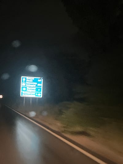 A blue directional signpost is visible at night on a wet road in Novi Iskar, Bulgaria. The sign has three sections, with text and arrows in white. The top section indicates "Plovdiv" with an upward arrow and the number "147". The middle section shows "Svilengrad" with an upward arrow and the number "234". The bottom section has an arrow pointing right for "Sofia" and the number "1". The road appears to be illuminated by headlights, reflecting on the wet surface. Trees and foliage are visible in the dark background. The image is blurry due to low light conditions and potentially movement.