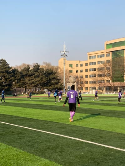 An outdoor soccer field in Taihe District, China, is depicted under clear skies with bright sunlight casting long shadows. The field is covered with artificial green turf marked with white lines. In the immediate foreground, a person is seen from behind, wearing a purple soccer jersey with the number "6" and the Chinese characters "马赫克" (Mahek) emblazoned on the back, black shorts, and light purple socks with yellow athletic shoes. This individual is walking towards the center-right of the frame.

Approximately ten to fifteen other individuals, primarily males in athletic attire including blue and purple jerseys, are scattered across the field, participating in what appears to be a soccer game or practice. A soccer ball is visible in the mid-ground. To the left of the field, a dense line of evergreen trees is present. In the background, a large multi-story building with a light-colored facade and numerous windows, some featuring green reflective glass, is visible. A tall stadium light pole with multiple floodlights stands prominently in front of the building. Bare deciduous trees are situated between the light pole and the background building. A portion of a soccer goal structure is visible on the far right edge of the field.