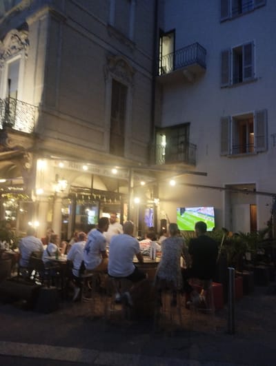 The image depicts an outdoor seating area of a restaurant in Lugano, Switzerland, likely in the evening or at dusk. A group of people are seated at tables and on stools, with some appearing to be watching a large outdoor television screen displaying a soccer match. The restaurant has an awning with lights, and the building facade is visible in the background. A street or sidewalk is in the foreground, with some potted plants and planters. The overall atmosphere suggests a casual dining or viewing experience.