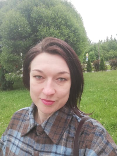 A medium shot selfie shows a woman with dark brown hair and fair skin standing outdoors. She is wearing a brown and blue plaid shirt and a dark brown strap across her shoulder. The background consists of lush green grass, a large tree, and several smaller evergreen trees. A traffic sign is visible in the distance. The lighting suggests an overcast day. The location is the city of Tosno, Russia.