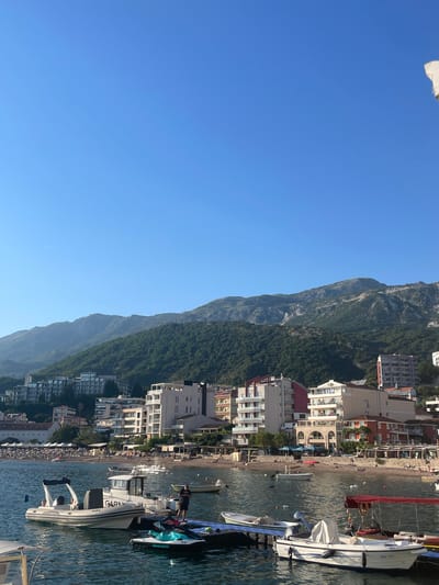 The image captures a coastal scene in Bečići, Montenegro, featuring a combination of natural and urban elements. The sky is a clear, gradient blue, taking up a significant portion of the frame. A towering, green, densely vegetated mountain range forms a vertical barrier along the right, extending into the background. The town of Bečići is built at the base of the mountains, visible as a series of buildings with varying heights and architectural styles. These structures include both residential and commercial properties.
The shoreline contains a sandy beach crowded with people. The beach transitions into the turquoise water of the Adriatic Sea, where several boats are moored. The boats in the foreground, near a pier, range from small vessels to larger yachts. A speedboat labelled with "GARMIN" is prominent near the bottom right of the frame.