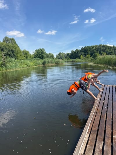 The image depicts a natural outdoor scene with two individuals dressed in orange life vests, in the act of diving off a wooden dock into a body of water. The water appears calm, with ripples visible on its surface. Lush green vegetation, including trees and reeds, lines the far bank of the waterway. The sky above is blue with scattered white clouds. The scene is consistent with a recreational area along a river or lake. There is no specific visual information in the image to confirm or deny its location within the city of Brest, Belarus.