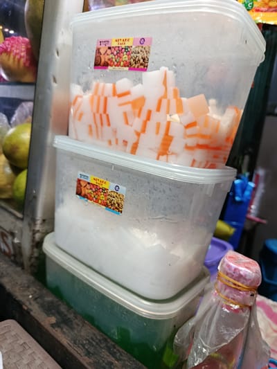 A close-up shot shows two stacked plastic containers filled with food items, possibly dessert or beverage ingredients. The top container is filled with diced white and orange cubes, while the bottom container holds a mass of white shreds. Both containers have condensation on their sides. A sticker on the top container displays the text "MIYAKO PACK" and images of nuts and candies. Another similar sticker is visible on the lower container. In the background, glimpses of other food items and containers are visible, including what appears to be a pink-frosted pastry and some green fruits. To the right, a bottle with a pink cap is wrapped in plastic and secured with yellow rubber bands. The scene appears to be at a food stall or market, likely in Depok, Indonesia, given the location context. The lighting suggests it might be daytime, but the overall setting is somewhat dim.