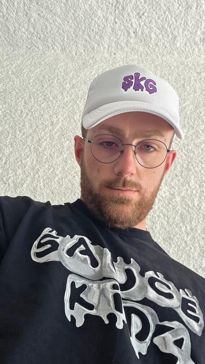 A man is pictured from the chest up, looking directly at the camera. He is wearing a white baseball cap with purple lettering that reads "SKG". He also wears round-rimmed glasses and a black t-shirt with a graphic design in shades of grey. The background is a textured white wall. The image does not provide any information about the city of Razlog, Bulgaria.