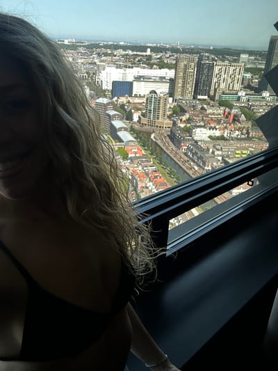 The image captures a person's head and shoulder, looking out over a sprawling cityscape from a high vantage point. The perspective is somewhat skewed, as the cityscape is oriented vertically. The scene depicts a dense urban environment with a mix of residential and commercial buildings, some modern high-rises, and what appears to be a river or canal running through the city. The sky is a clear, pale blue, suggesting daytime and fair weather.
In the foreground, the person's blonde hair is loosely styled and cascades around their face and shoulders. They are wearing a dark-colored top, possibly a bikini top, given the context of a swimwear suggestion. A delicate bracelet is visible on their wrist. The person's face is partially obscured by their hair and the angle of the shot, but they appear to be gazing out at the view.
The overall impression is one of leisure and enjoyment, with the person taking in a panoramic view of the city. The setting suggests an observation deck or a high-rise apartment, offering a commanding perspective. The urban landscape suggests a location like The Hague, Netherlands, known for its blend of modern architecture and historic districts. The time of day is clearly daytime, indicated by the bright natural light and clear sky. There is no visible text in the image.