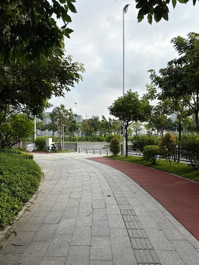 The image depicts a paved pathway in a park-like setting within Tianhe District, China. The pathway is made of light gray granite tiles with a textured tactile paving section for visually impaired individuals. Adjacent to the pathway is a red running track. Lush green bushes and trees line the path, with some trees bearing sparse leaves. In the background, modern buildings and construction cranes are visible, suggesting an urban environment. A lamppost stands tall in the distance. A white scooter is parked on the grass near the pathway. The sky is overcast with clouds. The overall scene conveys a sense of an urban green space designed for recreation.