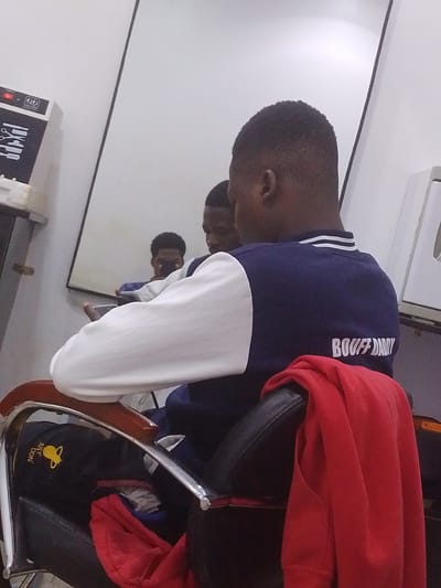The image depicts a young Black man sitting in a barber chair, seen from behind. He is wearing a navy blue varsity jacket with white sleeves and white and blue striped trim around the collar. White text on the back of the jacket reads "BOUFF DADY." A red sweatshirt is draped over the back of the chair. In the reflection of a large mirror behind him, another young Black man is visible, appearing to be holding a phone. The setting appears to be a barber shop, with a sterilizer device on a shelf to the left, displaying icons of scissors and combs. The lighting is artificial, suggesting an indoor environment, possibly late afternoon or evening. The overall mood is casual and relaxed. The location is identified as Abeere, Nigeria.