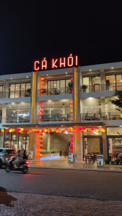 At night, a three-story building stands with bright interior lighting, decorated with hanging lanterns. A vertical neon sign reads "CA KHOI" in red letters. The building's facade is modern, featuring large windows and white pillars. A street runs alongside the building, with a motorcycle and a car visible on the pavement. The location is Quy Nhon Ward, Vietnam, indicated by the Vietnamese sign.