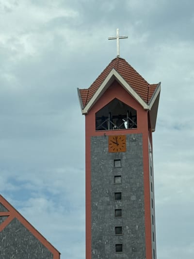 The image shows the bell tower of the Church of St. John Vladimir in Bjeliši, Montenegro. The tower is tall and square, with a pointed roof topped by a cross. The main part of the tower is red, with a gray stone facade. There is a clock on the tower, and several small windows. The sky is cloudy. In the foreground, a portion of the church roof is visible.