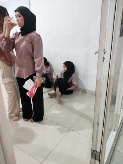 The image shows four women standing and sitting in a room, possibly an office or home. The women are all wearing hijabs, a headscarf worn by Muslim women. Three of the women are wearing pink long-sleeved shirts, and one woman is wearing a black long-sleeved shirt. One woman is sitting on the floor, while the other three women are standing nearby. One woman is holding a cup of a yellow liquid and drinking from it. Another woman is holding a red and white package with the word "Pretty" printed on it. The floor is tiled. The walls are white. There is a closed door to the right of the image. The coordinates -6.308, 107.282 locate a place in Telukjambe, Indonesia.