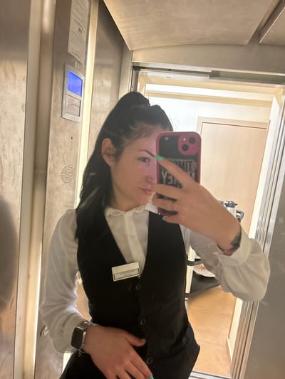 A woman is taking a selfie in what appears to be an elevator. She is wearing a white long-sleeved shirt, a black vest, and a name tag on her vest that reads "Maikela Kostadinova". She is holding a pink phone in her left hand. The elevator has metal panels and a control panel visible.
This location information could not be accurately assessed from the image.