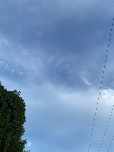 The image depicts a clear blue sky with scattered clouds, suggestive of a daytime scene. Three dark, thin lines, likely power lines, stretch horizontally across the upper portion of the frame, indicating an outdoor setting likely near human infrastructure. The lower right corner of the image is dominated by the lush green foliage of a large tree, its branches and leaves creating a dense canopy. The light and cloud patterns suggest it is a bright day, possibly late morning or early afternoon. The vibrant green of the tree indicates a healthy, well-watered environment. The scene evokes a sense of calm and natural beauty, with the man-made elements of the power lines contrasting with the organic presence of the tree and sky. The location context of Lafia, Nigeria, suggests a tropical or subtropical climate, which aligns with the appearance of the dense, green tree. No people or specific events are depicted, and there is no visible text in the image.