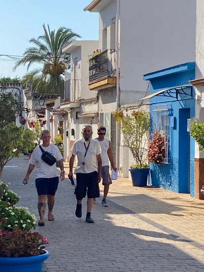 The image captures a sunny day in Cancelada, Spain, focusing on a narrow, cobblestone street lined with white buildings. Three individuals are walking away from the camera. The person in the foreground, on the left, is wearing a white t-shirt, dark shorts, and a black fanny pack. They have short, blonde hair and appear to be looking forward. Next to them, slightly behind and to the right, is another person in a white t-shirt and dark shorts, with a visible beard. This individual is also looking forward and holding what appears to be a booklet or papers. Further to the right and slightly behind them is a third person in a white t-shirt and grey shorts, wearing sunglasses and also looking forward.
The buildings are characteristic of Mediterranean architecture, with balconies adorned with potted plants. The street is illuminated by bright sunlight, casting distinct shadows across the cobblestones. Potted plants and flowers are visible along the sides of the street, adding color to the scene. A prominent blue building with white-trimmed windows is on the right side of the image. A palm tree is visible in the upper left portion of the frame, indicating a warm climate. The overall impression is of a pedestrian street in a pleasant, sunny environment.