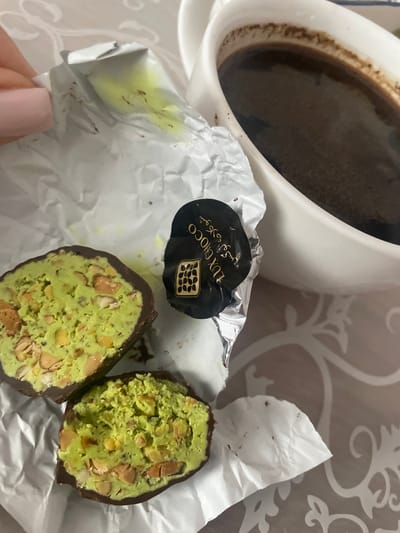 In this close-up, top-down shot, a white mug of dark coffee sits on a patterned tablecloth, likely in a home setting in Brovary, Ukraine. The coffee appears to be freshly brewed, with a fine layer of grounds visible along the rim of the mug. Next to the mug, a piece of silver foil wrapper, some of which is crumpled and torn, holds two halves of a chocolate-covered confection. The confection has a dark chocolate shell and a vibrant green filling studded with nuts, possibly pistachios, lending it a textured, somewhat rustic appearance. The wrapper itself shows some specks of brown, suggesting chocolate debris.
A person's hand, with a pale pink fingernail, is visible in the lower left corner of the image, gently holding a section of the wrapper. The overall impression is one of a casual, perhaps indulgent, moment. The lighting suggests it could be daytime, though no direct sunlight is visible. The embossed pattern on the tablecloth, featuring stylized floral or vine motifs in a lighter shade of beige, adds a touch of subtle elegance to the scene. One of the wrapper pieces has black text printed on it, which reads "LUX CHOCO" and some Arabic script, possibly indicating the brand or flavor of the chocolate. The image captures a simple pleasure – coffee and a sweet treat.