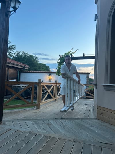 A man in a white shirt and shorts stands on a wooden deck, leaning on a railing. He is looking towards the camera. The deck is surrounded by a wooden fence. In the background, there is a white wall with a light fixture and some greenery. The sky is blue with some clouds. The image appears to have been taken in the city of Voyvodino, Bulgaria.