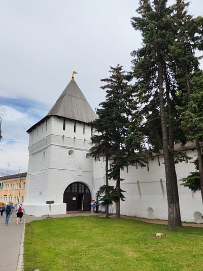The image depicts the Spaso-Preobrazhensky Monastery in the city of Shebunino, Russia. A prominent white brick tower with a wooden conical roof topped by a golden flag stands on the left. The tower has an arched wooden entrance. To the right of the tower is a long white wall, also made of brick. Several tall evergreen trees with dense foliage are positioned in front of the wall and partially obscure it. A grassy area with a small tree stump is in the foreground, to the right of a paved walkway. On the walkway, a few people are visible. Two individuals walk away from the camera on the left, one with a blue backpack and another with a red backpack. To the right of the tower's entrance, a small group of people stands, appearing to be interacting. In the background, to the left of the tower, a building with yellow and white facades and several windows is visible, suggesting an urban environment adjacent to the monastery. The sky is overcast with some visible clouds.