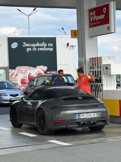 This image was taken at a Shell gas station in Drazhevo, Bulgaria. In the foreground, a dark grey Porsche 911 Carrera GTS is parked at a pump. The license plate is PB 2206 XE. A man in an orange and yellow uniform, likely a gas station attendant, is holding a fuel nozzle and appears to be speaking with the driver of a dark blue SUV, who is partially visible through the Porsche's open window. Another man wearing glasses is standing next to the SUV. In the background, a large billboard features a "Shell Café" logo and text in Bulgarian that translates to "You deserve a break." Several cars are present at the gas station, including a silver Mazda in the far left. The sky is overcast with some clouds.