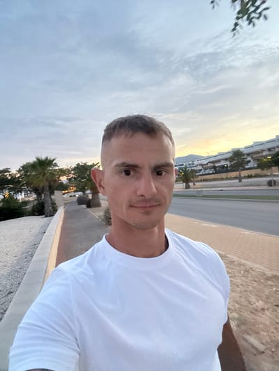An outdoor, eye-level, medium shot selfie shows a fair-skinned man with short, dark hair and a buzz cut. He is wearing a white t-shirt and is looking directly at the camera. The man is standing on a paved walkway in Finestrat, Spain. To the left of the walkway is a gravelly area with a low concrete wall. Behind the wall, there are palm trees and foliage. Across the street from the walkway are modern buildings. The sky above is cloudy, with a hint of yellow from the sunset. The overall scene appears to be a residential or resort area.