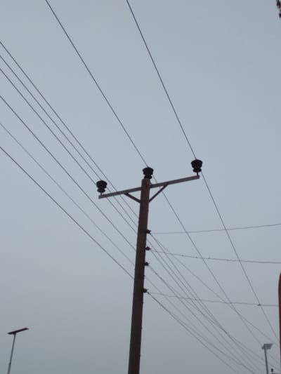 The image presents a view of a utility pole and an intricate network of overhead power lines set against a uniformly overcast, light gray sky, consistent with an outdoor scene in Ejule, Nigeria.

A prominent utility pole, likely made of wood or concrete, stands vertically in the mid-right portion of the frame. Near its top, a horizontal cross-arm extends, supporting three distinct black, spool-like insulators. From these insulators, three main power lines extend across the upper part of the frame, stretching diagonally from left to right.

Numerous other, thinner electrical wires crisscross the sky, both connected to the main pole at various points (including smaller insulators lower down) and extending out of the frame in multiple directions, creating a dense web. This suggests a complex electrical distribution system.

In the bottom-left corner, the top of a modern streetlight or similar tall pole is partially visible. A shorter pole, possibly another streetlight, is also faintly discernible in the bottom-right background. The consistent grayness of the sky indicates an overcast day, suggesting diffused light conditions without direct sunlight. There are no people visible, and no specific activities or events are taking place. No text is visible anywhere in the image.