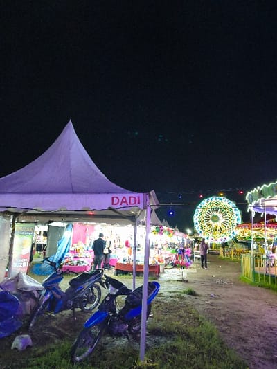 The image depicts an outdoor night market or fairground scene in An Khe Ward, Vietnam. The time is nighttime, characterized by a dark, clear sky above artificially lit structures.
In the foreground, on the left, stands a white pyramidal tent with the word "DADI" inscribed in red capital letters on its front canopy. Directly in front of this tent, two blue motorcycles are parked on a ground composed of dark soil with patches of green grass and some visible debris. Within the "DADI" tent, merchandise, appearing to include toys and other retail items, is displayed on tables. Two individuals are discernible inside this tent.
A dirt path extends from the foreground into the background, flanked by numerous other vendor stalls and tents, all emitting light. In the mid-ground, towards the right, a large, brightly illuminated, circular structure, resembling either a Ferris wheel or a decorative light display, is prominent with multi-colored lights. To its immediate right, a partial view of another illuminated carnival ride, potentially a carousel, is visible behind a green fence. Several other human figures are present in the mid-ground and background, appearing to walk along the path or stand near the stalls. The overall environment is bustling with activity and illuminated by various artificial light sources emanating from the market stalls and attractions.