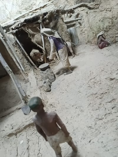 The media file shows a scene with two apparent human figures, likely mannequins or dolls, in a dimly lit, rustic setting. One figure is lying down, partially covered by a burlap-like material and what appears to be fabric clothing in shades of purple and white. It is positioned in what looks like a shallow pit or crevice within the earthy surroundings. The other figure is standing, facing away from the camera, and is also composed of a dark material with lighter patches, possibly representing skin or clothing. Its head is topped with a dark green object.
The ground is uneven and appears to be made of dirt and straw, with some debris scattered around. There are hints of rough wooden structures and possibly some discarded objects like a piece of tire in the background. The overall impression is of an abandoned or neglected space.
The lighting is subdued, suggesting it might be indoors or outdoors at dusk or dawn, or in an area with limited light sources. There are no clear indicators of specific weather conditions. The environment suggests a rural or perhaps an undeveloped area, consistent with the provided location context of Angren, Uzbekistan, which is known for its industrial and mining activities, but also has rural surroundings.
The figures' poses and the general state of the scene do not convey any specific activity or event. They appear to be static. There is no visible text in the image. The emotion evoked by the scene is one of stillness, abandonment, and perhaps a touch of desolation due to the rough materials and dim lighting.