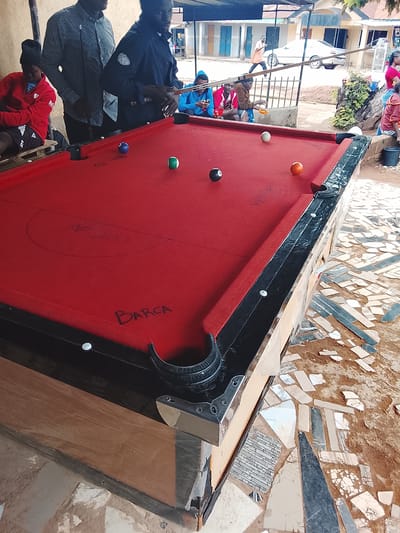 The image depicts an outdoor scene in Kabong, Nigeria, centered around a red felt billiards table. Several individuals are present. In the foreground, a portion of the billiards table is visible, with several balls scattered on its surface: a blue ball with a number "8", a white ball, a green and white striped ball, and an orange ball. The table has black rails and a wooden base. In the background, various people are observed. A person in a red jacket and black beanie sits on the left, looking away from the camera. To their right, two individuals stand behind the billiards table. One, wearing a blue jacket, appears to be playing pool, holding cue sticks. Another person, in a checkered shirt, stands beside them. Further in the background, a few more individuals are seated or standing, and a white car is visible on a street. The environment appears to be a semi-urban or roadside setting with buildings and some vegetation. The ground is covered with a mosaic of broken tiles and earth. The overall impression is a casual gathering centered around a game of billiards in a Nigerian community.