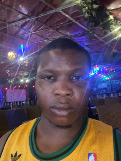 A man is facing the camera, illuminated by multicolored lights. He wears a yellow and green basketball jersey with an NBA logo. Behind him is a structure, possibly a decorative cage, adorned with more lights and greenery. His skin has a sheen, and his expression is neutral as he looks directly into the lens. The composition suggests an outdoor setting or a space decorated for an event or festivity.
