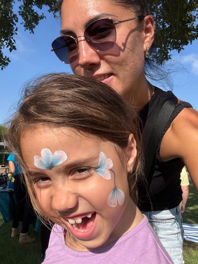 In Netanya, Israel, a young girl with brown hair and a purple shirt, her face adorned with light blue flower face paint, laughs joyfully as she poses for a selfie with a woman. The woman, wearing sunglasses and a black top, is positioned behind the girl, her arm around the child. The background reveals a sunny outdoor setting with a clear blue sky, green grass, and trees. In the distance, a table with various items and a person in a blue shirt can be seen, suggesting a public event or gathering.