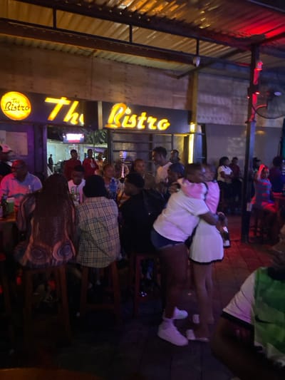The image shows the outdoor seating area of a bar called "The Ristro" in Dodoma, Tanzania. There are many people sitting at tables and chairs. The people are mostly young adults, wearing casual clothing. Some are talking, laughing, and enjoying drinks while others are hugging and interacting with each other. The bar has a wooden exterior and is lit up with bright yellow lights. The seating area is brightly lit, with a wooden floor and wooden tables and chairs. There is a ceiling fan hanging overhead. The environment is lively and friendly.