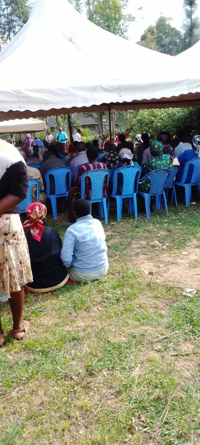 A large white tent is erected over a gathering of people seated in blue plastic chairs. The tent is supported by a metal frame and has a white canvas covering. The people are seated in rows facing a speaker. There are a total of 30 people in the image, most of whom are women.  The people are seated on the ground and there is grass and dirt in front of the chairs. Some of the people are wearing traditional clothing. The event is taking place in a grassy area in Kipkarren, Kenya. The coordinates are 0.599 latitude and 34.957 longitude. It appears to be a church service or gathering as there are several people wearing religious clothing. The event is taking place in an outdoor environment with trees and a roof in the background. The atmosphere is peaceful and serene.