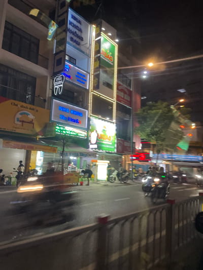 This is a night-time, vertical-oriented photograph of a street scene in Thủ Đức, Vietnam. The image is somewhat blurry and appears to be taken from a moving vehicle. The scene depicts a street lined with multi-story buildings, many of which have illuminated signs.
The subjects include several motorbikes parked along the street and in front of the buildings. Some people are visible on or near the motorbikes. The buildings house various businesses, indicated by the illuminated signs. One sign reads "NHA KHOA SKY DENT'S" in blue letters with a white outline, suggesting a dental clinic. Another sign has green lettering and a circular logo. A yellow sign with red and blue text is also visible.
The time of day is clearly night, with artificial lights from signs and street lamps illuminating the scene. The weather is not explicitly discernible but the lack of rain suggests it might be clear. The presence of motorbikes and the style of the buildings and signage are strong indicators of a location in Vietnam. The overall atmosphere is that of a busy commercial street at night. The blurriness of the image might convey a sense of movement and the dynamic nature of urban life.