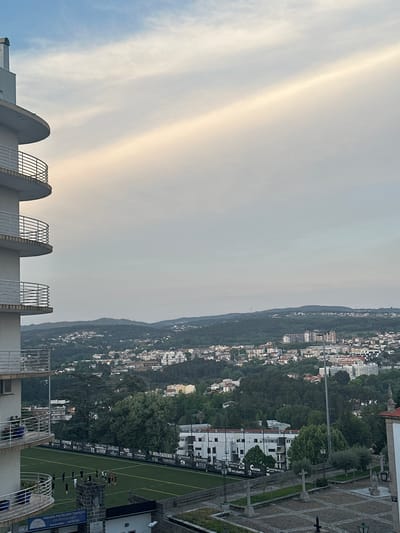 The image depicts a cityscape of Vila Real, Portugal, under a sky with diffuse sunlight. The foreground shows a partial view of a building with architectural elements including concrete arcs and metal fencing. In the middle ground, a green sports field with players practicing is visible. Advertisements border the field. Beyond the field lies a densely populated hillside with buildings interspersed among trees. In the upper portion of the image, the sky presents a hazy appearance with subtle color variations. The city sprawls over the landscape. A sign advertises: Padaria Pastelaria Tradição Churrasqueira.