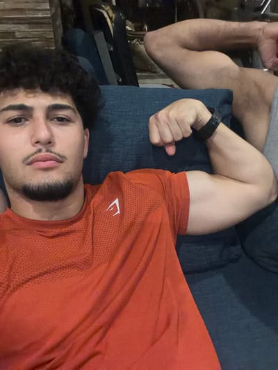 The image shows two men posing for a selfie. The man in the foreground is looking at the camera, with his face filling the left side of the frame. He has dark, curly hair, a mustache, and a goatee. He is wearing an orange t-shirt with a white logo. His right arm is flexed, and he is wearing a black watch on his left wrist. The man in the background is partially visible on the right side of the frame. He is also flexing his arm, and his bicep is prominent. They appear to be indoors, possibly in a living room or lounge area, as suggested by the dark blue couch and wooden background elements. The overall lighting is dim. The image does not provide any specific information to determine the location within Sarouja Municipality, Syria.