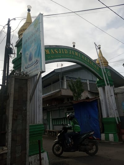 A mosque entrance, marked "MASJID JAMI AL-ISLAH," features a green archway topped with golden domes. A blue and white sign for "AIR MINUM Isi Ulang" (refillable drinking water) stands to the left. A motorcyclist wearing a green jacket is riding on the road in front of the entrance. The background shows a two-story building and a makeshift shelter covered with a blue tarp. Power lines stretch across the overcast sky.