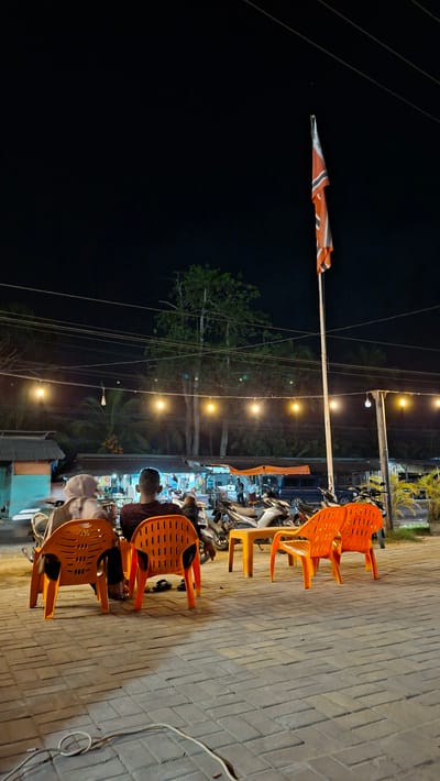 The image depicts an outdoor scene at night, possibly a roadside cafe or food stall, in Keudeteupinpunti, Indonesia. The foreground features several bright orange plastic chairs arranged on a brick-paved walkway. One chair is occupied by a person seen from behind, wearing a dark red patterned shirt, sitting at a table. Another person is partially visible in a chair further down.
Behind the chairs, a row of motorcycles is parked, suggesting a popular local spot. A string of yellow lights illuminates the area, creating a warm ambiance. Beyond the lights, trees and foliage are visible against the dark night sky. A tall pole with a flag or banner attached to it stands prominently on the left side of the frame. The flag has orange and white diagonal stripes. Electrical wires crisscross the scene overhead.
The overall impression is of a lively evening gathering, likely a place where people stop for a meal or a drink. The time of day is clearly night, indicated by the darkness and artificial lighting. The location cues, such as the motorcycles, the style of the chairs, and the tropical vegetation, suggest a warm climate. The scene appears casual and relaxed. No distinct text is visible in the image.