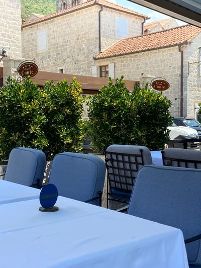 The image shows an outdoor seating area in front of a stone building with red tile roofs, likely in the city of Perast, Montenegro, as suggested by the "Hotel Conte" sign visible on the building. In the foreground, there are several tables covered with white tablecloths. One table has a blue oval "RESERVED" sign on it. Blue upholstered chairs with patterned cushions are placed around the tables. Behind the tables, there are lush green bushes with yellow-tinged leaves. The building behind the bushes is constructed of light-colored stone and has windows with white shutters. Two cars are visible parked on the street behind the bushes, and a portion of a mountain is visible in the upper left background. The scene is illuminated by daylight, suggesting it is daytime.