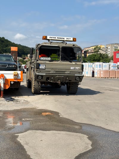 The image shows a military-style truck parked on an asphalt surface. The truck is olive drab and appears to be a recovery or utility vehicle. It has large, off-road tires and a robust chassis. A yellow light bar with the text "ПЪТНА ПОМОЩ 24/7" (Road Assistance 24/7) is mounted on the front. Below the windshield, there is a license plate that reads "EBBP 5078".

The truck is parked next to what appears to be a construction or industrial area, with stacks of building materials visible in the background. There are also buildings and trees in the distance, suggesting an urban or semi-urban setting. The ground is damp in places, indicating recent rain or a water spill. The sky is clear and blue, suggesting a sunny day. The overall mood of the image is utilitarian and functional, focusing on the vehicle and its purpose. The presence of the building materials and the vehicle's designation imply ongoing work or a service being provided.