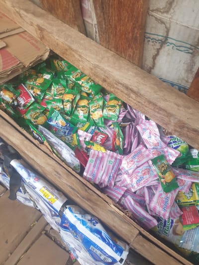 The image shows a wooden crate overflowing with various packaged candies and some cleaning detergent products. The candies are in individual wrappers, many of them in green packaging with the text "Candy Ginger" visible. There are also pink wrappers with the text "Splash Strawberry & Cream Candy". Some cleaning detergent packages, with blue and white branding, are visible at the bottom and left side of the crate. The background appears to be a rough, possibly wooden or cardboard wall. The lighting suggests an outdoor or dimly lit indoor environment. There are no people visible in the image. The scene is a close-up shot focusing on the contents of the crate. The origin and location of this image are not discernible from the visual information provided.
