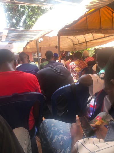 The scene depicts an outdoor gathering within the Akpaka Forest Reserve, Nigeria. Numerous individuals, predominantly male, are seated on blue plastic chairs under a light-colored tarp canopy that provides shade from visible overhead sunlight. All individuals face forward, oriented towards a presentation area. At the front, a person stands near a whiteboard displaying written content, appearing to address the assembled group. In the immediate foreground, a person is actively using a smartphone with both hands, while another individual holds a small printed item. Audience members are dressed in various casual attire.