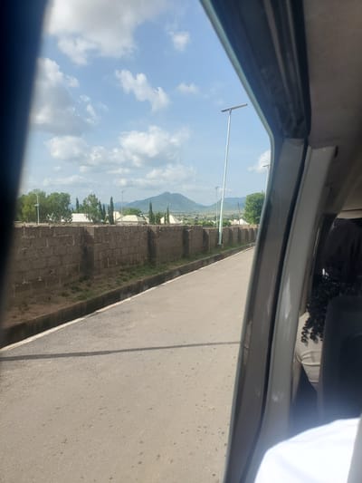 This image captures a view from inside a vehicle, looking out towards a road in the city of Kuson, Nigeria. The road is paved with asphalt and has a concrete drainage channel running alongside it. On the left side of the road, a tall, gray brick wall extends into the distance. Beyond the wall, there are several buildings with white roofs, trees, and tall, thin streetlights. In the background, a large, green mountain range is visible under a partly cloudy sky. The right side of the image shows the interior of the vehicle, including the door frame and a glimpse of a person's head and shoulders with dark, curly hair.