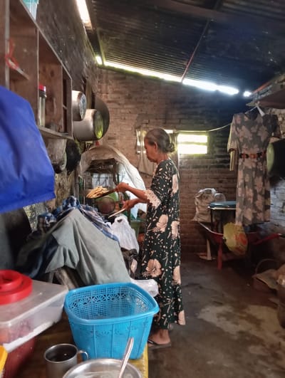 In a humble kitchen in Baron, Indonesia, an elderly woman with her hair pulled back in a bun stands focused as she uses a slotted spoon to transfer freshly fried food from a pan. She wears a dark green dress adorned with a delicate floral pattern and white lace on the cuffs. The kitchen itself is rustic, with exposed brick walls and a corrugated metal roof that allows shafts of sunlight to stream in, illuminating the dusty interior. Shelves made of wood are filled with various kitchenware, including pots and containers. In the foreground, a blue plastic basket sits on a table next to storage containers, one with a red lid. A wheelbarrow and sacks of what appear to be goods are visible in the background. The overall scene depicts a moment of everyday life and domestic activity in a simple, functional living space.