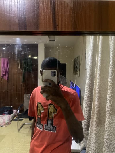 This media file depicts a mirror selfie taken by a person in a room. The subject is a young person with short, dark hair, wearing an orange t-shirt with the number "11" printed in gold and black on the front. The t-shirt is the focal point of the image, with the "11" design prominently displayed.
The setting appears to be a residential interior, possibly a bedroom or living area. In the reflection, we can see a beige curtain with a decorative pattern, a computer monitor displaying a blue screen, and some objects on a shelf. There's also a glimpse of wooden furniture on the left side of the frame, and what looks like a purple garment hanging in the background. The lighting suggests it is daytime, though the exact time is not discernible. The room appears to be somewhat cluttered, with a chair and some items on the floor visible in the reflection. The image has a casual and personal feel, characteristic of a selfie. No specific emotions are conveyed by the subject. There is no visible text in the image other than the number on the t-shirt. The location context is Giginyu, Nigeria.