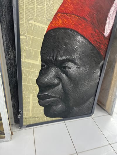 This image captures a framed artwork resting on a white tiled floor in an indoor setting within Alaguntan, Nigeria. The artwork, a textured painting or mixed-media piece, depicts the left profile of a dark-skinned individual, likely an adult male, with a neutral facial expression. The figure wears a prominent red cap, textured with visible brushstrokes, which includes a subtle white feather-like embellishment on its right side. The background of the artwork consists of a collage of light-toned, aged paper sections, appearing as columns of printed text. An inscription, "Adegoke Nurudeen. M 4", is visible in the lower-left section of the artwork's background. The piece is contained within a dark grey or black frame. Other partially visible framed artworks or canvases are present on both the left and right sides of the main subject, leaning vertically against an unseen surface. The floor beneath consists of white square tiles with dark grey grout lines.
