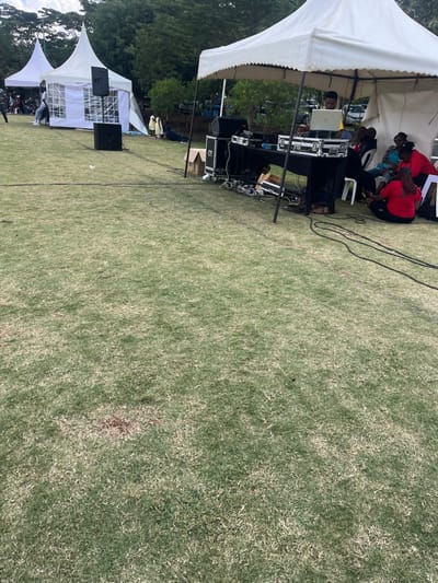 An outdoor event features white tents on a grassy field. People are gathered under the tents. Audio equipment and a DJ setup are visible in one tent. The grass covers most of the frame, with some tree coverage on the left side. Power cords run across the grass.